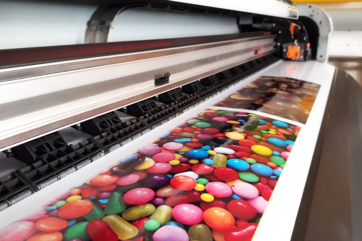 A wide-format digital printer meticulously applying colorful inks onto a roll of printable vinyl. A large format printer laying down ink on a roll of printable vinyl